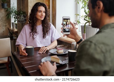 Young man taking photo of his girlfriend sitting by table in front of him and posing while having coffee with yummy dessert - Powered by Shutterstock