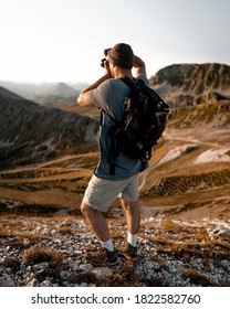 young man taking a photograph of sunrise on top of a mountain, unmarked clothing with backpack hat and trekking boots