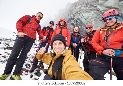 Young Man Taking Picture With Friends Hikers During Travel In Winter Mountains. Group Of Male Travelers Looking At Camera And Smiling, Showing Thumbs Up. Concept Of Travelling, Hiking And Photography.