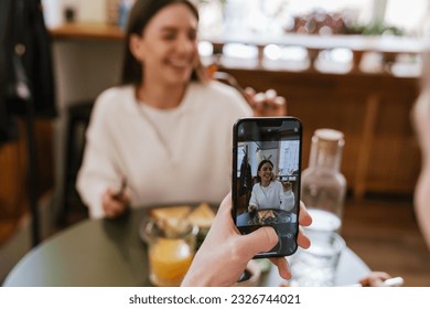 Young man taking pictures of his smiling girlfriend with mobile phone while having lunch together in cozy cafe - Powered by Shutterstock