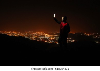 Young Man Taking Selfie On Top Of The Hill Observing The Night City View.