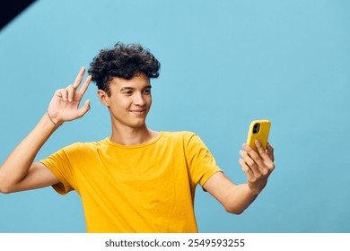 Young man taking a selfie with a smartphone against a light blue background, smiling and making a peace sign, showcasing modern technology and social interaction - Powered by Shutterstock