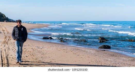 Young Man Is Taking A Walk On The Beach. He Is Sad And Thoughtful.