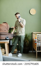 Young Man Talking On Mobile Phone While Caring About His Newborn Baby In Cradle At Home