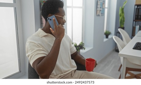 Young man talking on phone holding red mug in modern office interior with large windows and plants - Powered by Shutterstock