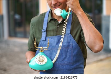 Young Man Talking On The Vintage, Old Phone Standing Outdoors Wearing Apron. Service Man Having Conversation Using Old Phone. Telephone Communications Concept. No Face Visible. 