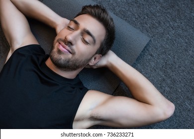 Young man in tanktop lying on a yoga mat with eyes closed and hands behind head