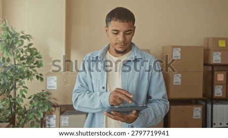 Young man tapping tablet with hands among stacked cardboard boxes in a small e commerce packing area inside a building; entrepreneurial confidence.