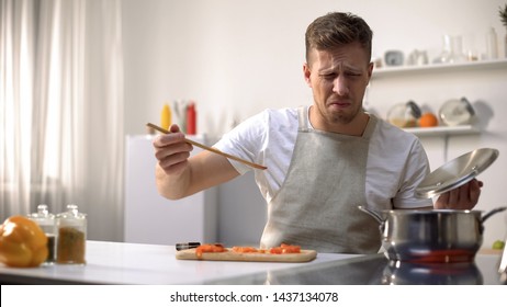 Young man tasting cooked food with disgusted face expression, funny grimacing