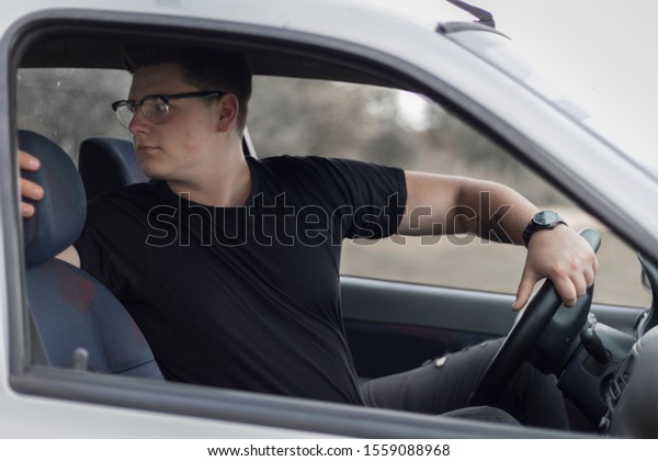 Young man with tattoo in\
black t-shirt sitting in car and holding steering wheel and looking\
back