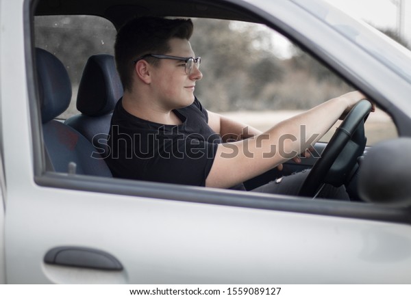 Young man with tattoo in black\
t-shirt sitting in car and holding steering wheel and\
driving