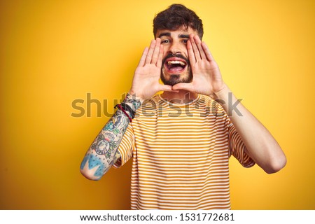 Young man with tattoo wearing striped t-shirt standing over isolated yellow background Smiling cheerful playing peek a boo with hands showing face. Surprised and exited
