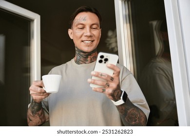 A young man with tattoos smiles as he sips coffee and looks at his phone on a cozy morning. - Powered by Shutterstock