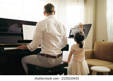 The Young Man Teacher Have Online Piano Lesson Music Class At Home Which Has A Little Daughter Girl Kid Nearby.