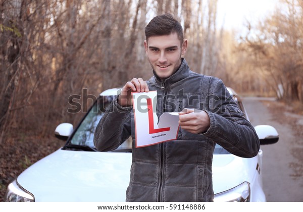 Young man tearing learner driver sign while\
standing near car\
outdoors
