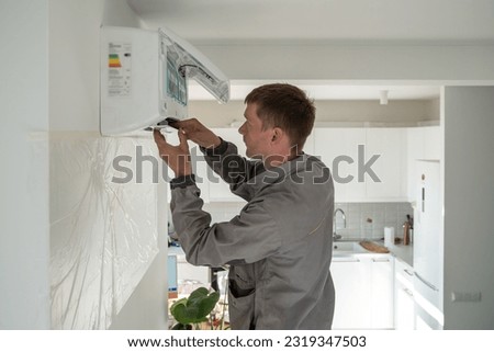 Young man technician worker in uniform fixing repairing apartment air conditioner, installing wall-mounted mini split. AC unit maintenance concept
