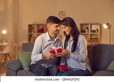 Young Man Tenderly Looks At His Girlfriend Giving Her A Gift For Valentine's Day. Loving Couple Exchanging Gifts Sitting On The Couch At Home In A Cozy Room. Valentine's Day Concept.