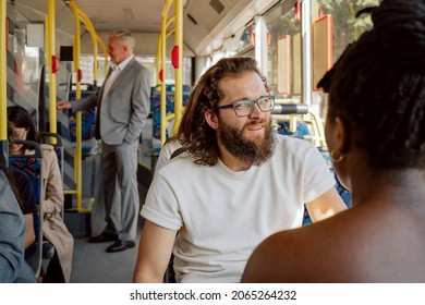 Young man with thick beard long hair wearing glasses sits on public transport bus in front of dark-skinned colleague with dreadlocks tied up in bun friends returning from work college talking laughing