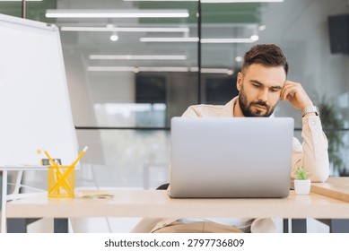 Young man thinking while working on laptop, sitting at desk in an office workspace, focusing on business tasks