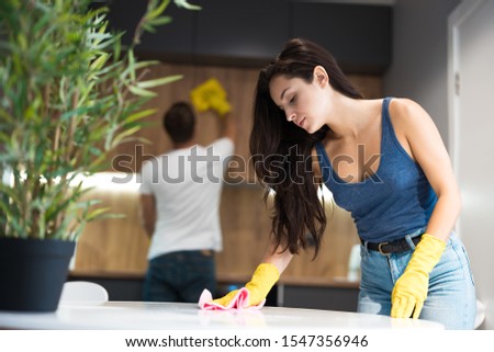 young man thoroughly wiping dust off from the kitchen cabinet with a rag meanwhile beautiful woman wiping kitchen table in yellow gloves doing cleaning together