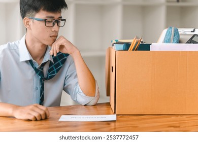 A young man thoughtfully surveys a box of materials and papers on a table, suggesting contemplation or transition in a workspace. - Powered by Shutterstock