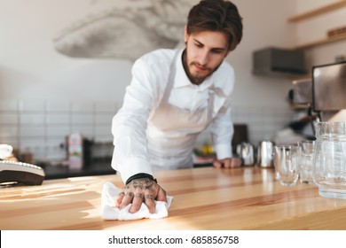 Young man thoughtfully wiping counter at his workplace in restaurant. Portrait of barista in apron and white shirt working at the counter in coffee shop. Young barista cleaning counter at cafe - Powered by Shutterstock