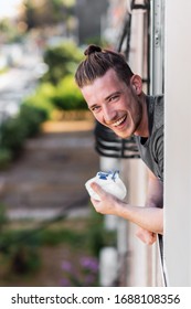 Young Man Took Off His Medical Mask And Looks Out Of The Apartment Window, Smiles At His Neighbor And Hopes For A Quick End To Quarantine. Taken During The Coronavirus Epidemic. New Reality