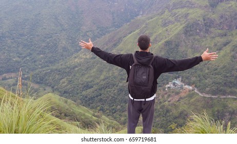 Young Man Tourist With Backpack Standing On The Edge Of Beautiful Canyon And Victoriously Raised Hands. Male Hiker Reaching Up Top Of Mountain And Outstretching Arms Up. Rear Back View.