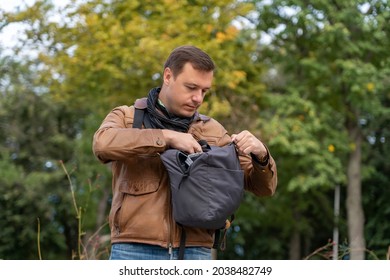 Young Man Tourist Standing, Searching And Getting Something Out Of His Backpack In Autumn Park Outdoors. Portrait Of Millenial Man Holding Backpack At Street. . Travel Luggage And Packing Concept