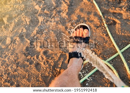 Young man tourist washing bare foot in summer sandal with hose water jet on sand Prasonisi beach under sunset light in Greece