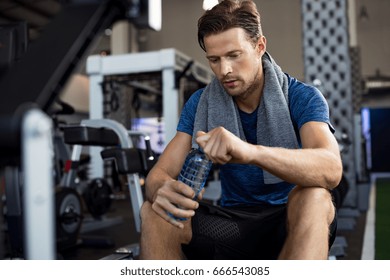 Young man with towel on his shoulders drinking water while sitting at gym. Sweaty guy opening cap of bottle to drink water after workout. Tired man resting after training and holding bottle of water. - Powered by Shutterstock