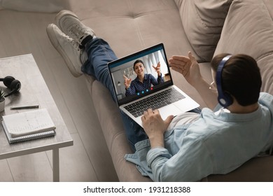 Young man trainee sit on couch in wireless headphones listen to indian woman trainer on pc screen. Focused guy student meet with biracial female teacher on remote consultation. Over shoulder top view