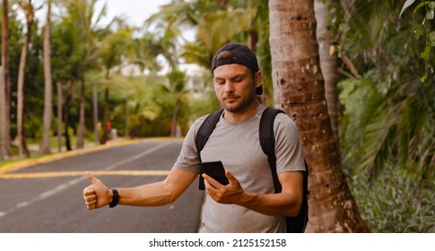 Young Man Traveler In A Cap With A Backpack Stands On The Road And Looks At The Screen Of A Mobile Phone In His Hand. Hitchhiking, Thumb Up, Waiting For A Taxi, Tourism, Travel Concept.