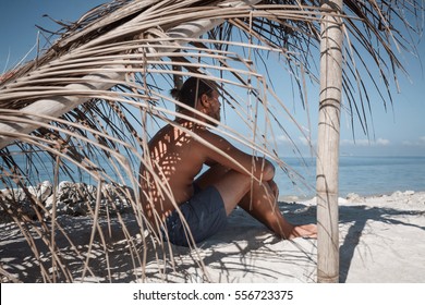 Young Man Traveler Sitting On Beach In The Shade Of Hovel Made Of Palm Leaves