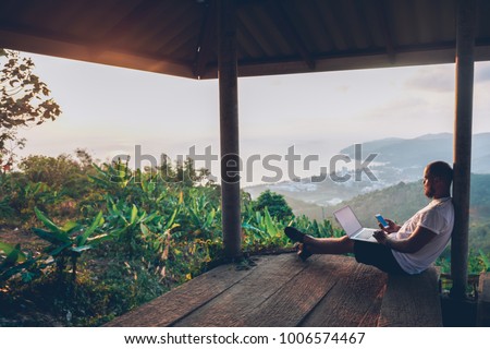 Young man traveler using laptop computer while enjoying beautiful tropical sunset in Thailand. Freelancer guy working on freelance on netbook during vacation holidays in summer. Male adventure blogger