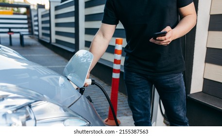 Young man traveling by electric car at summer, having stop at charging station standing plugging cable and browsing internet on smartphone joyful while charing