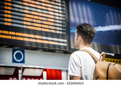 Young man traveling, reading big electronic train timetable in railway station, seen from behind