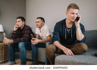 Young Man Trying To Talk On The Phone. Behind Friends Are Watching TV. Group Of Friends Watching A Football Match On TV Against The Background Of Young Guy Phone Conversation.