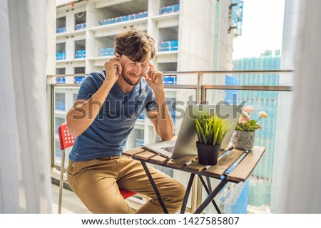 Young man trying to work on the balcony annoyed by the building works outside. Noise concept. Air pollution from building dust