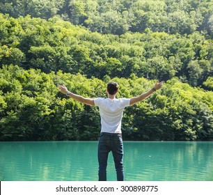 Young man in t-shirt, outdoor with arms spread open enjoying freedom in front of lake, seen from the back.