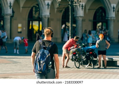 A Young Man In A T-shirt With A Travel Backpack On His Back And Headphones On His Head Travels Through The Old Town In Front Of A Man And A Woman Traveling On Bicycles, A Sunny Day In Krakow