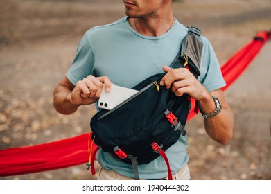 A Young Man In A T-shirt And A Waist Bag On His Shoulder Puts A Smartphone In His Pocket, Close Up