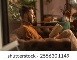 Young man in turtleneck sweater reading book at cozy cafe, side profile through window