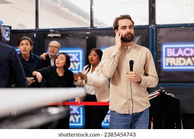 Young man TV news reporter standing with microphone against angry crazy crowd of shoppers waiting for discounts. Journalist correspondent making special reportage about Black Friday event