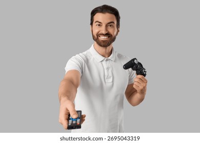 Young man with TV remote controller and game pad on light background