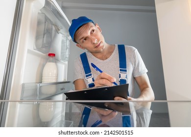 Young man in uniform writes down the refrigerator breakdowns with a pen on paper. Concept of repair work, the call of the master of repair of household appliances. Photo from inside the refrigerator
