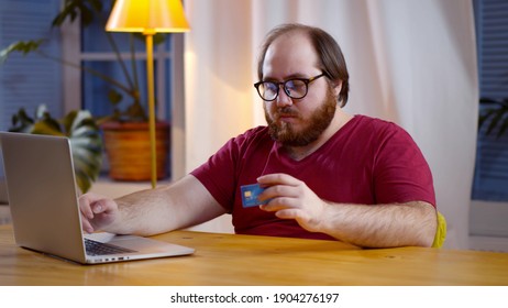 Young Man Use Credit Card For Shopping Payment Online On Laptop. Portrait Of Overweight Bearded Guy Sitting At Table And Paying Bills Online Using Computer And Credit Card