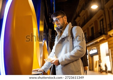 Young man using an automated teller machine, holding euro banknotes in a city street