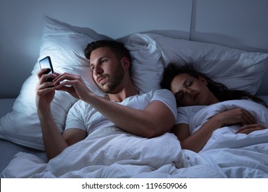Young Man Using Cellphone While Her Wife Sitting On Bed At Night