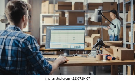 Young Man Using Desktop Computer with a Dashboard and Orders Information Display. Distribution Manager and Employee Working in Distribution Warehouse with Cardboard Boxes. Over the Shoulder Footage.